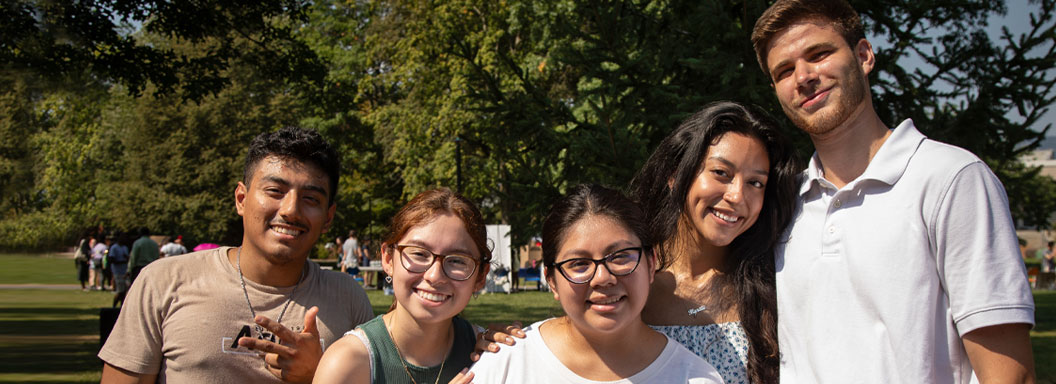 Two male and three female students smile outside on a sunny day at SUNY New Paltz.
