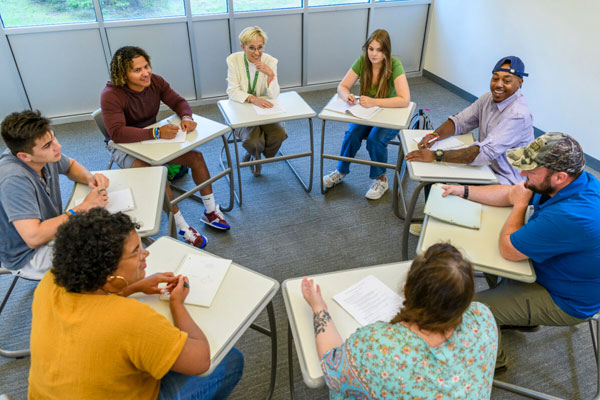 Eight people sit in school desks aligned in a circle for a meeting.