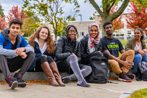 Students sit on the curb of a sidewalk for group photo with jackets on and fall trees behind them.